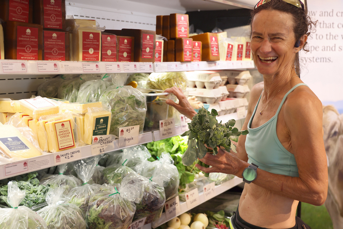 laughing woman holding broccoli in front of display cooler full of produce and cheese