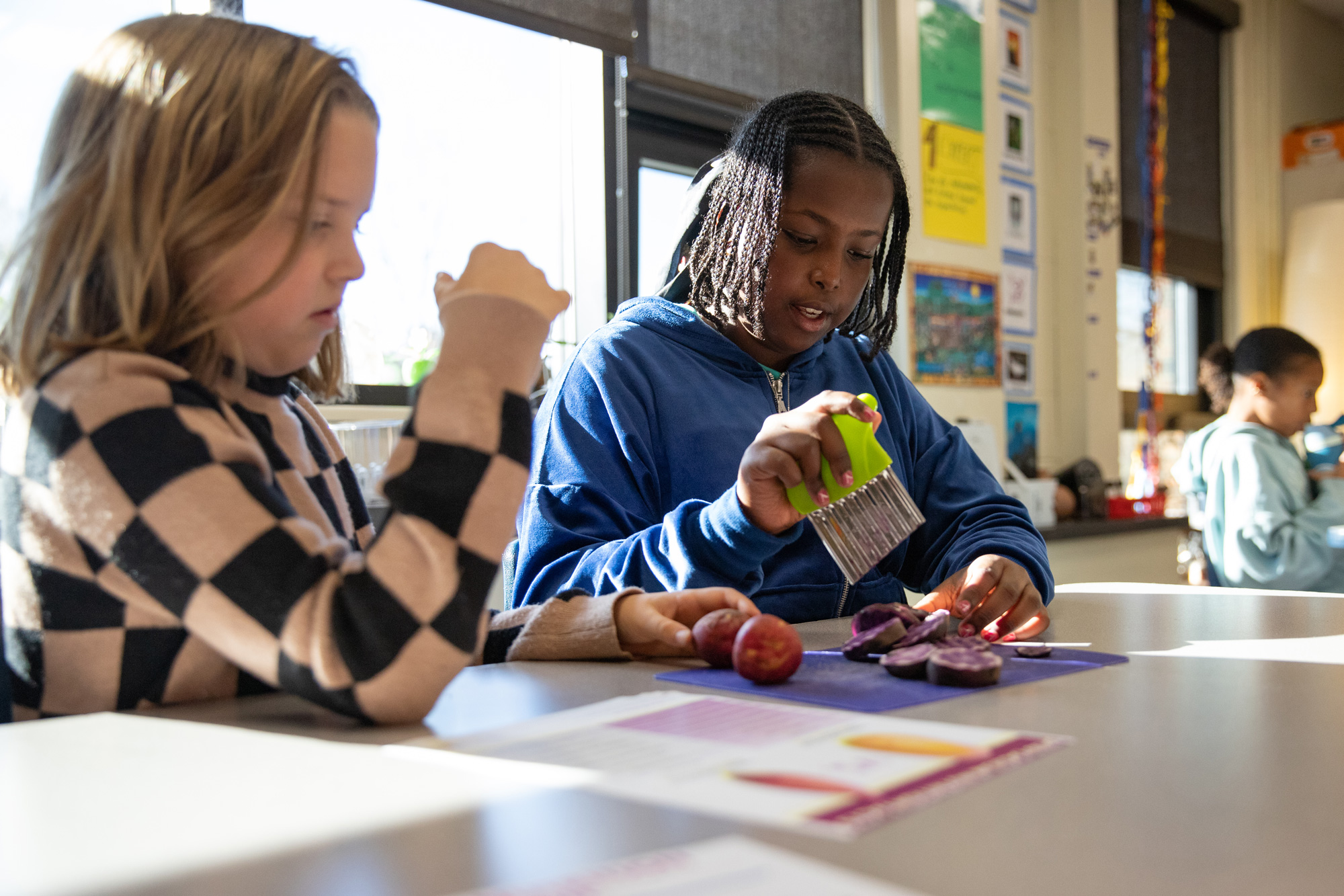 Two students at a desk in a classroom, working on a cooking project with potatoes.