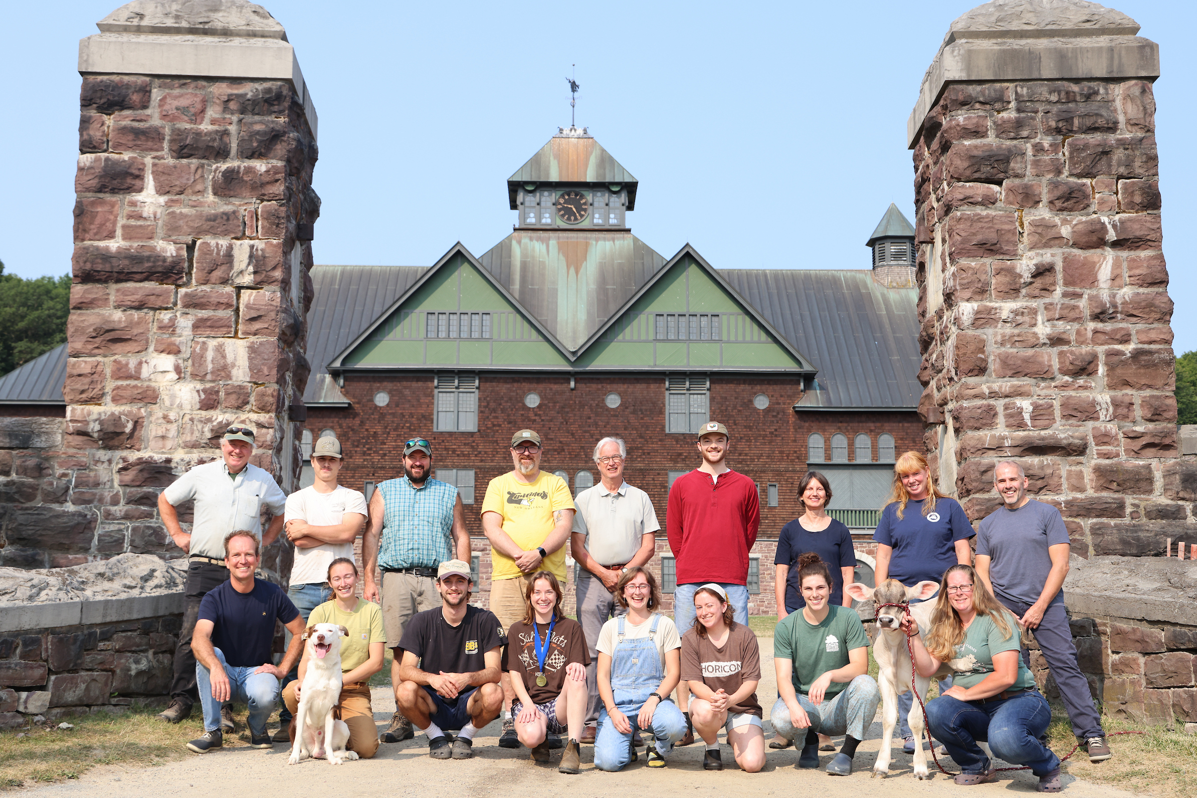 8 people kneeling between stone pillars on dirt entrance to big shingled building. Woman far left has a dog; woman at far right has a calf. Back row of 9 people standing.