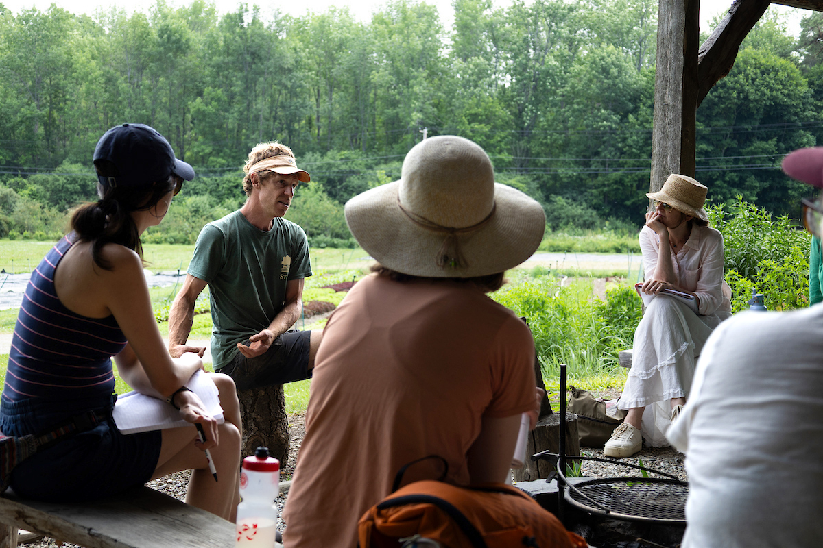 people sitting on benches in a circle, under an open air pavilion, backs to camera, listening to man talk