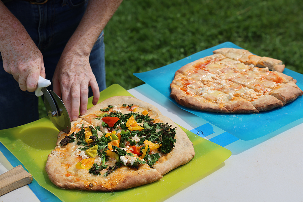 Two fresh pizzas sit on cutting boards being sliced for serving with a pizza cutter