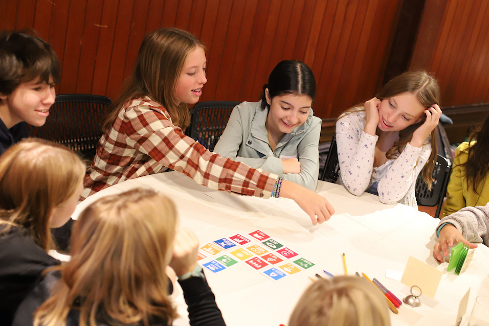 young teens sitting at a white tablecloth table displaying cards representing the U.N. Sustainable Development Goals. One girl is reaching over table for one of the cards