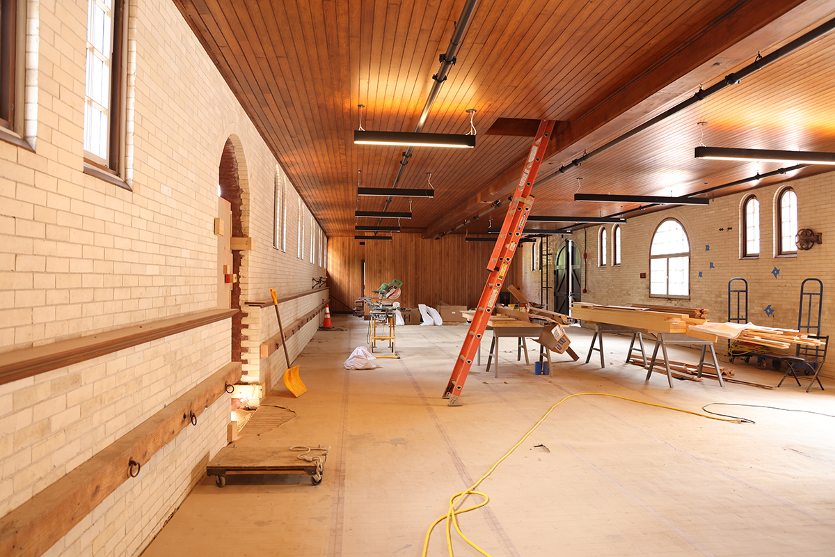 long big room under construction with white brick walls, arched windows, and construction equipment around, including a tall orange extension ladder leaning upright against a part of the wooden beadboard ceiling