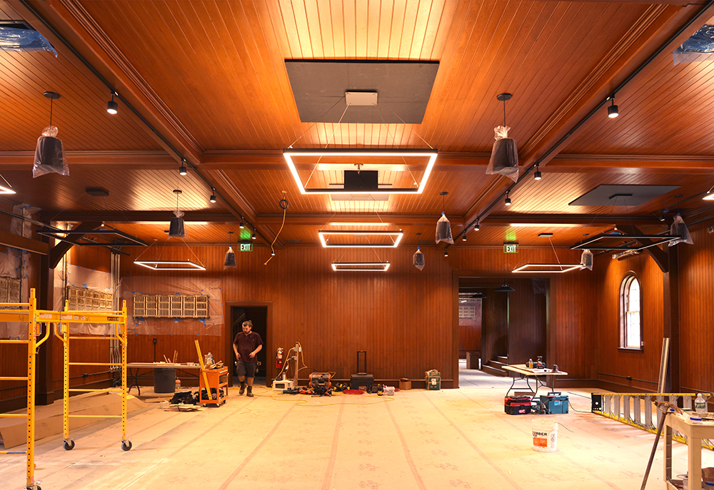 room under construction with staging, ladders on floor, tools, and protective material on floor. Man walking through door at back of the room