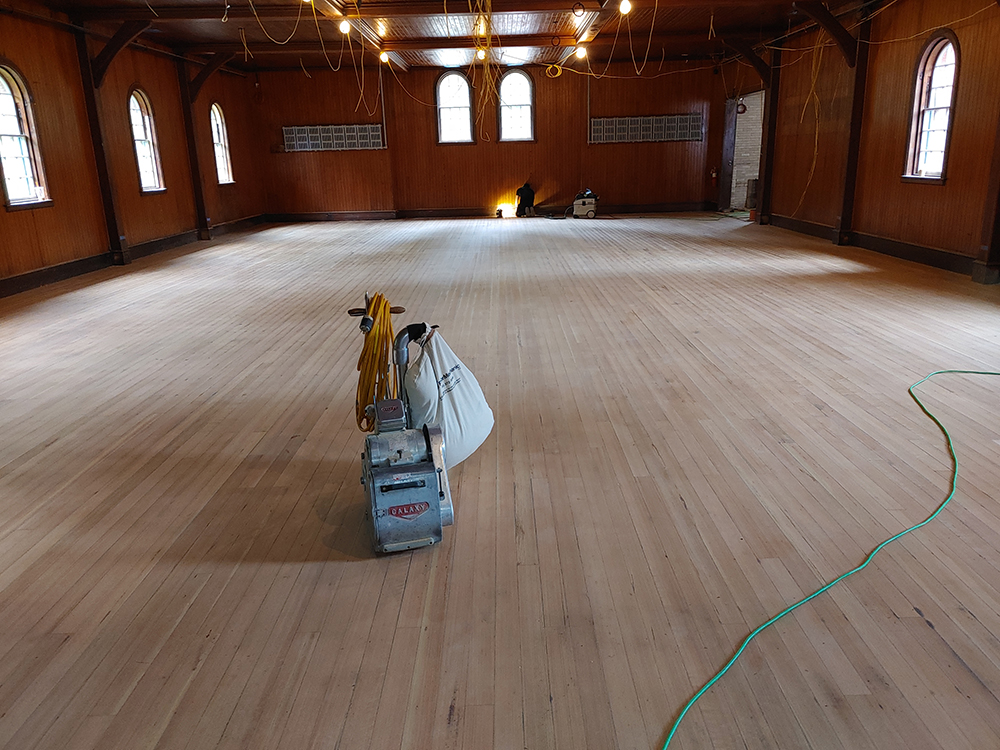 big room with paneled walls and recently sanded wooden floor. A floor sander is in the middle of the photo