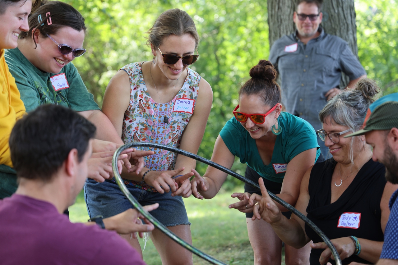 Seven people smile in a circle while balancing a hula hoop on their hands during a team building activity