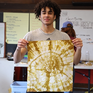 A teenage boy poses with a tie dyed bandana in a classroom