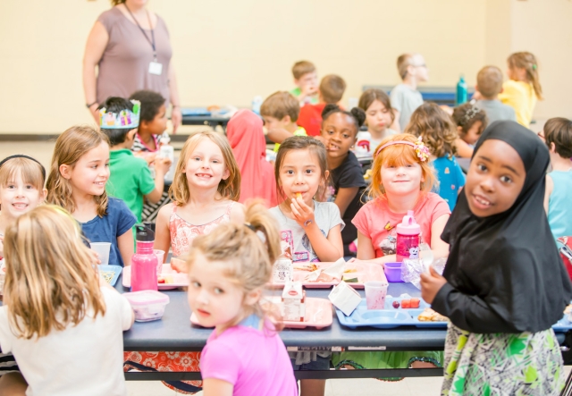 Group of children enjoying lunch together at school cafeteria tables, some looking at the camera and smiling.
