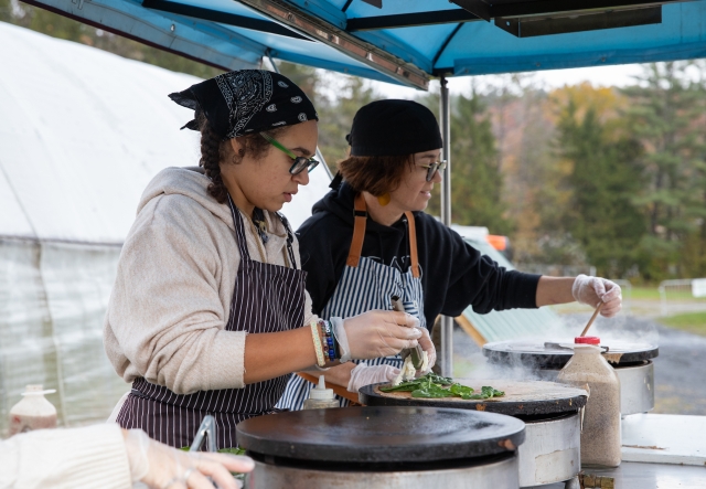 Two people in striped aprons are cooking at an outdoor food cart.