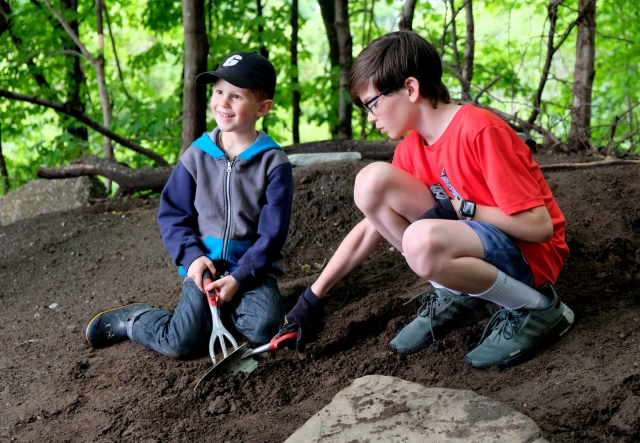 Two young children smile while digging with small trowels in a dirt patch