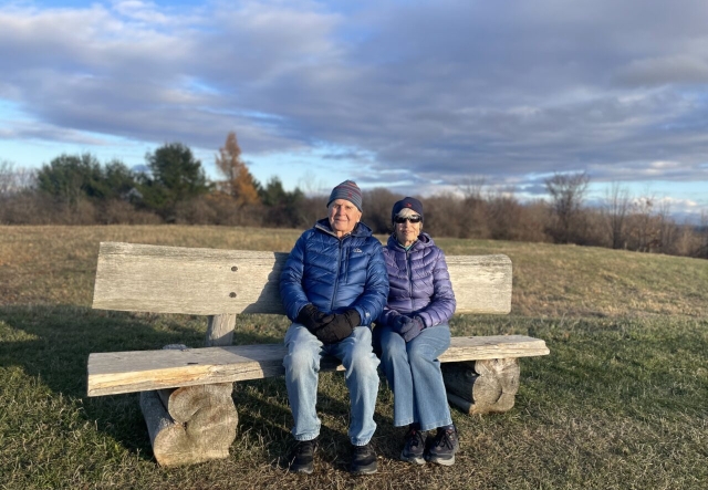 an older man and woman sit together on a rustic wooden bench surrounded by autumn grass. They are both in jeans, wearing winter coats, hats, and mittens.