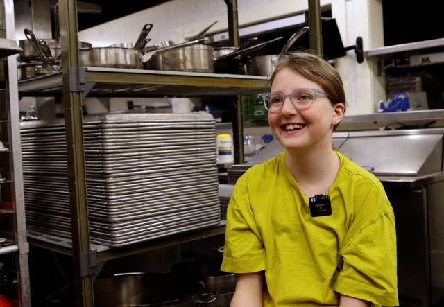Child in a green shirt and glasses smiles at the camera in a professional kitchen setting.