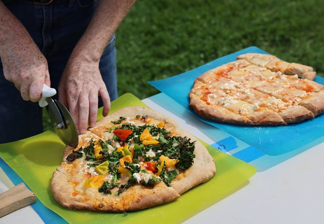 Two pizzas sit on cutting boards, one is being cut by a person with a pizza cutter