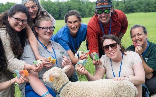 A group of people smiling, gathered around a lamb. They are holding colorful felted figurines.