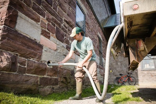 A man holds a hose, coming from the tail end of a truck, up through a circular hole carved from a stone wall.
