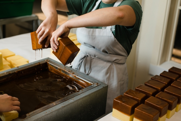 A worker pulls two blocks of cheddar, one in each hand, out of a vat of wax as excess wax drips off.