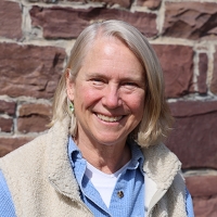 Woman with short, light blonde hair smiles at the camera inf front of a brick wall in an outdoor setting.