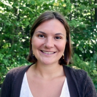 Person with shoulder-length brown hair smiles at the camera with green foliage in the background.
