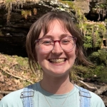 A young woman with light brown hair and glasses smiles at the camera in a natural setting.