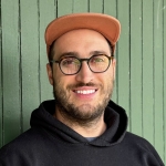 Bearded man in glasses and orange rimmed hat smiles at the camera in front of a green paneled wall.
