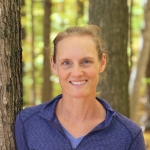 woman in blue shirt with blond hair tied back stands beside a tree in a forest of yellow autumn leaves
