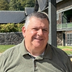 man with short graying hair in an olive collared shirt smiling at camera.