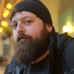 young man with bushy brown beard and mustache and wearing a black cap.